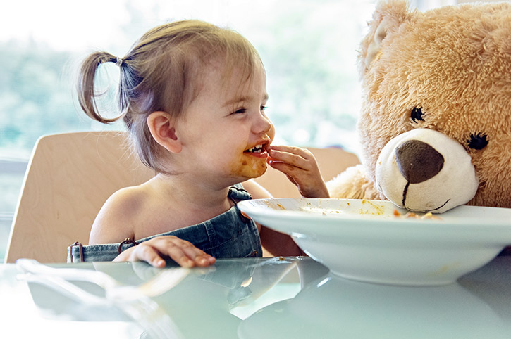 Pequeña comiendo con las manos ensuciando su boca acompañada de su oso café de peluche